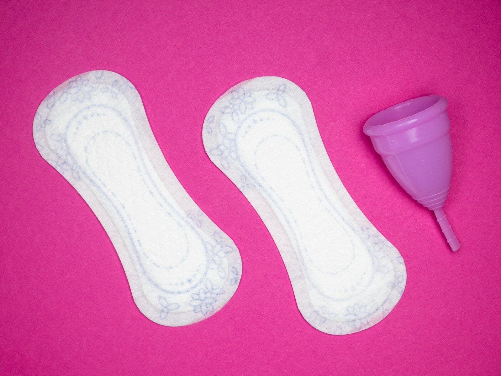 Photo by Reproductive Health Supplies Coalition a pair of menstrual pads next to a menstrual cup on a hot pink backgrouund. Naturopathic care for PMS can make your time of the month more bearable